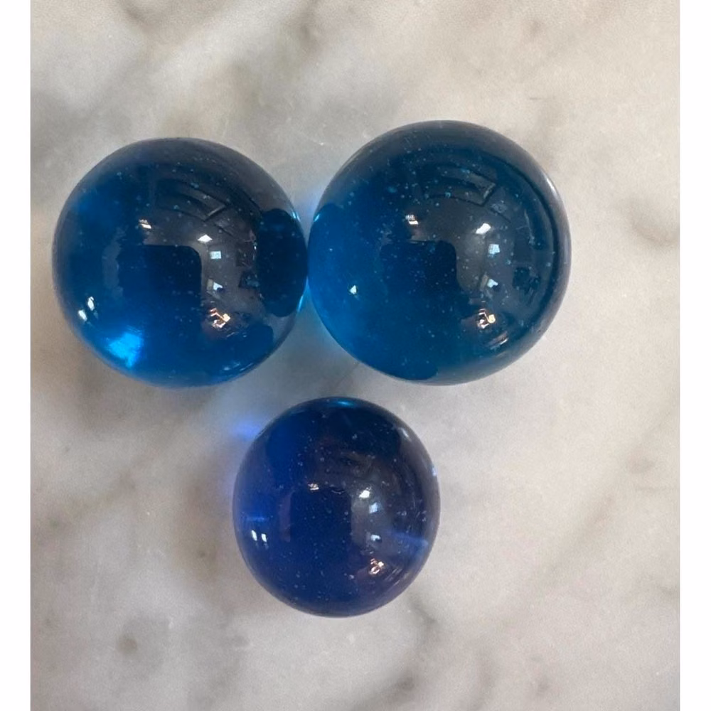 translucent blue glass marbles studio photograph - VTG Shooters Blue Glass Marbles Set of 3 Translucent Teal Cobalt Spheres 22-25mm Set of three vintage glass marbles features a striking translucent blue color with subtle internal bubbles and a smooth, polished finish. These spheres catch the light beautifully, making them ideal for decorative displays, floral arrangements, or as part of a classic marble collection. The set includes two slightly larger marbles and one smaller marble, all crafted from high-quality solid glass with a consistent deep teal and cobalt hue. Measurements: • Large Marble Diameter: 25mm • Small Marble Diameter: 22mm Material: Glass Color: Blue, Teal, Cobalt Quantity: 3 Shape: Sphere Finish: Polished, Translucent Style: Vintage, Classic Pattern: Solid with internal bubbles Transparency: High Theme: Play, Decor, Coll